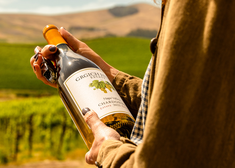 A woman is marveling at the bottle of Napa Valley Chardonnay in her hands before opening it with a corkscrew. An expanse of regenerative organic vineyards rolls in the background.