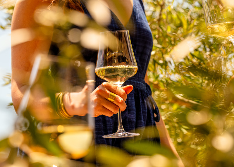 A woman in an olive grove, a glass of white wine in her hand. There is a second glass of white wine in the foreground.