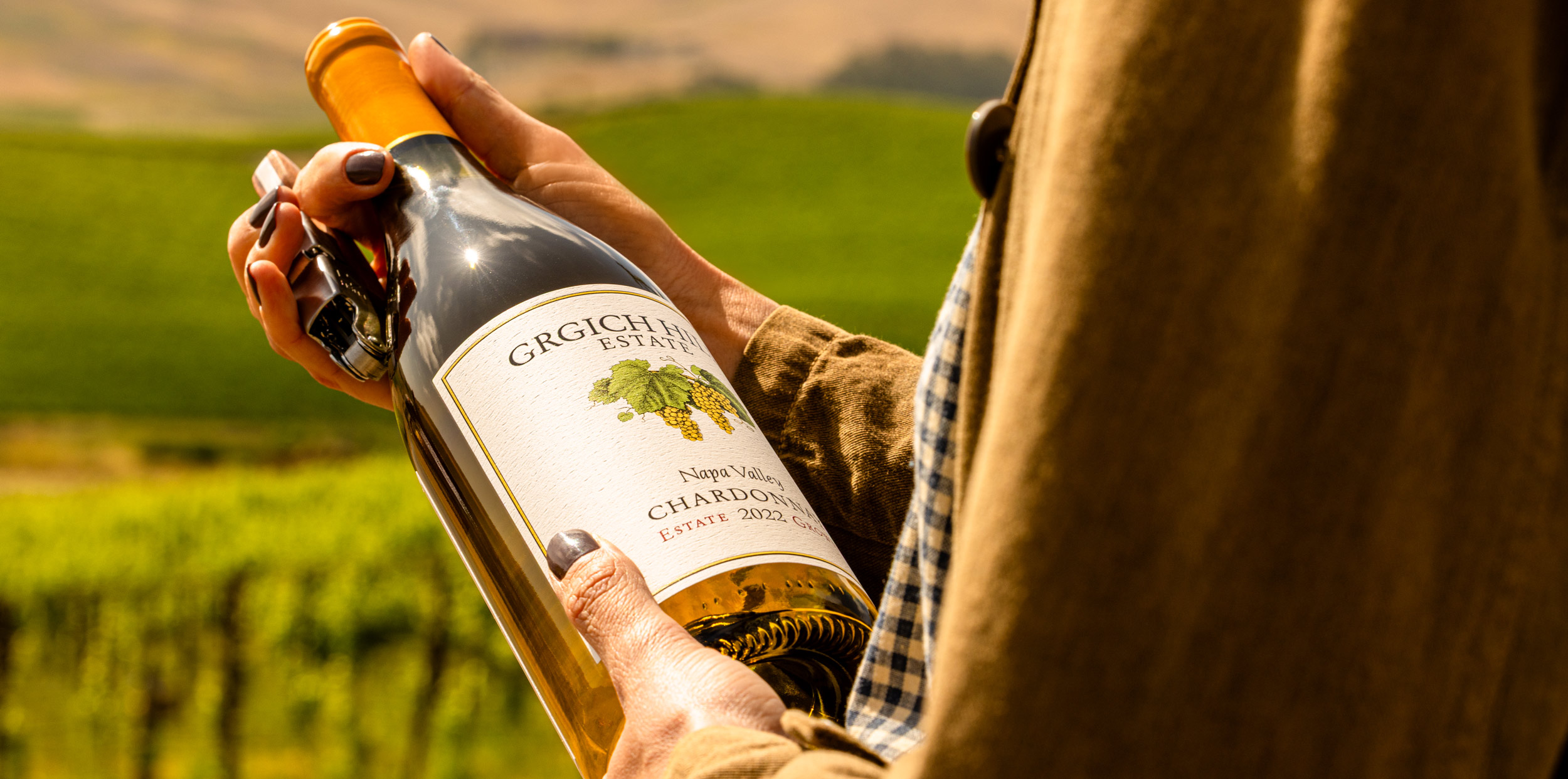 A woman is marveling at the bottle of Napa Valley Chardonnay in her hands before opening it with a corkscrew. An expanse of regenerative organic vineyards rolls in the background.