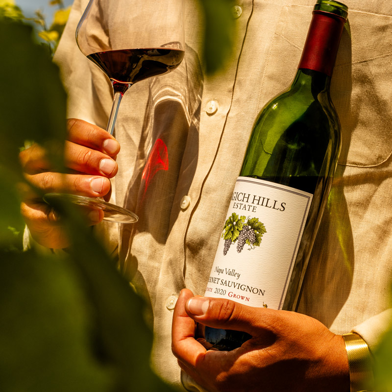 A young man cradles a bottle of Napa Valley Cabernet Sauvignon from Grgich Hills Estate. In his other hand, a glass of red wine makes red light dance across his shirt.