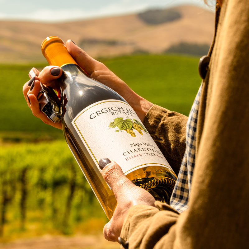 A woman is marveling at the bottle of Napa Valley Chardonnay in her hands before opening it with a corkscrew. An expanse of regenerative organic vineyards rolls in the background.