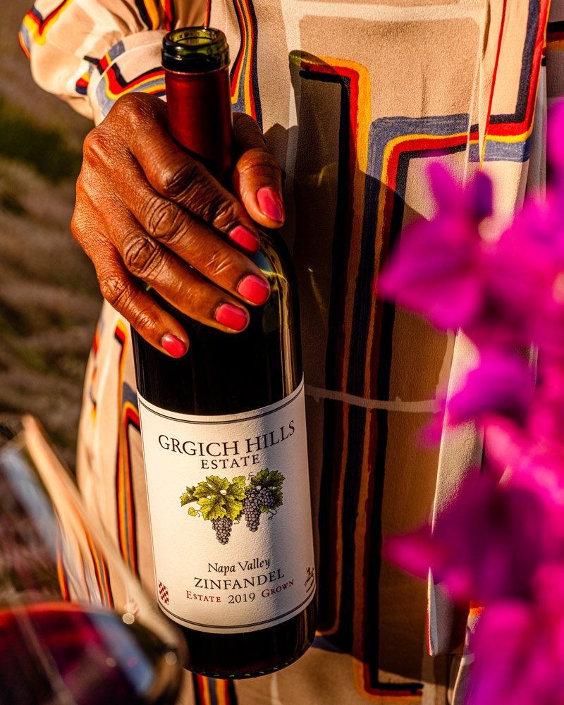 A hand holds an open bottle of Napa Valley Zinfandel from Grgich Hills Estate, with vibrant magenta bougainvillea in the foreground.