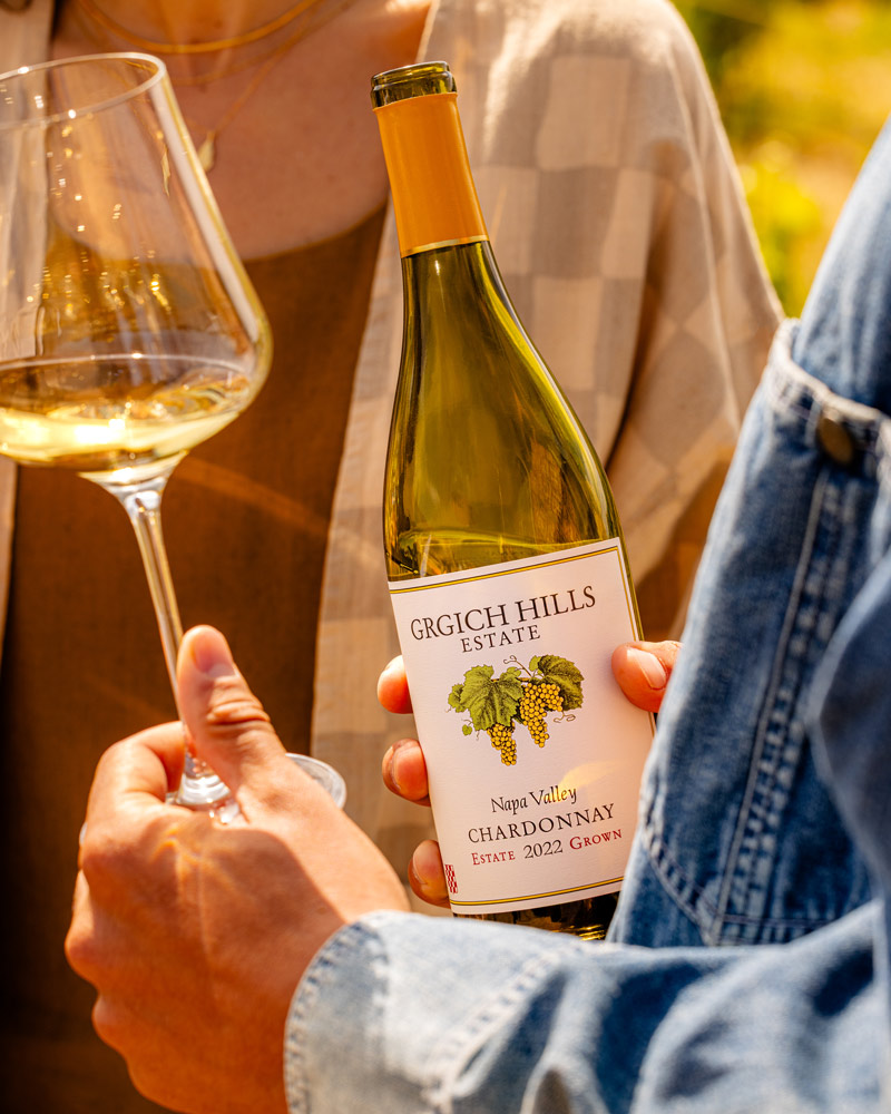 A young man is holding an open bottle of Grgich Hills Estate Chardonnay from Napa Valley in one hand. In his other hand, he swirls around a glass of Chardonnay.