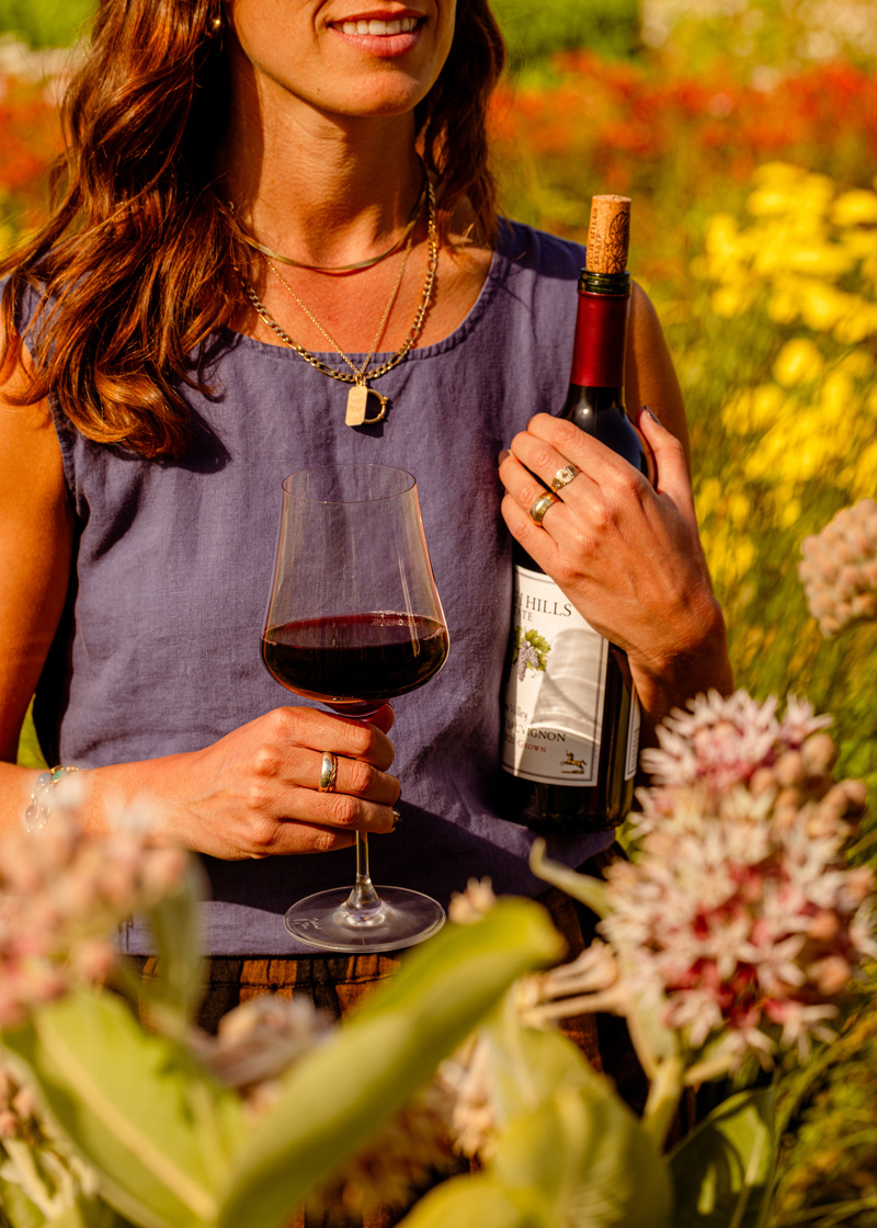 A young woman holds a bottle of Napa Valley Cabernet Sauvignon close while strolling through a field of flowers in a regenerative pollinator habitat.