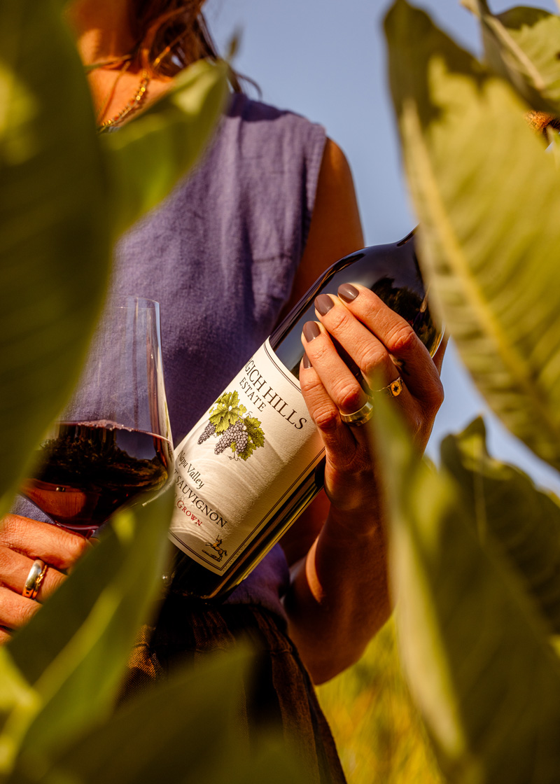 A hand clutches on a bottle of Napa Valley Cabernet Sauvignon, viewed through the parting of leaves.