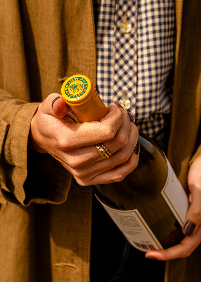 A bottle of Grgich Hills Estate's Napa Valley white wine is held gingerly in manicured hands, with the iconic grape cluster logo adorned on the foil.