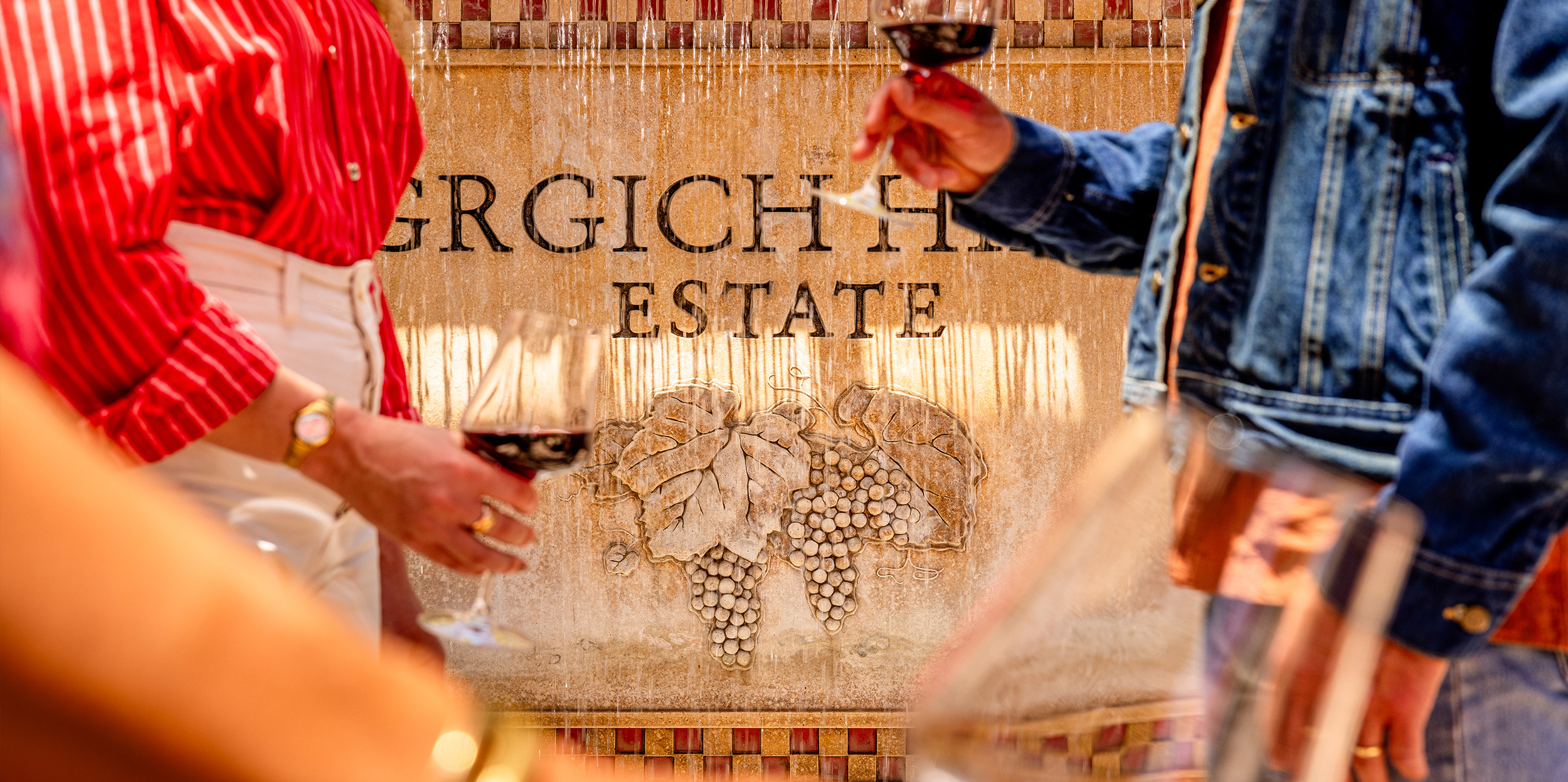 Two people enjoy a glass of Napa Valley Cabernet Sauvignon in front of a fountain bearing the Grgich Hills Estate logo and signature grape cluster.