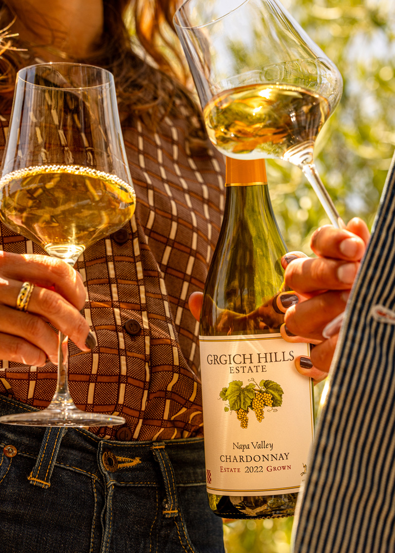 A young woman is holding an open bottle of Grgich Hills Estate Chardonnay from Napa Valley in one hand. In her other hand, she swirls around a glass of Chardonnay, standing across from a young man doing the same.