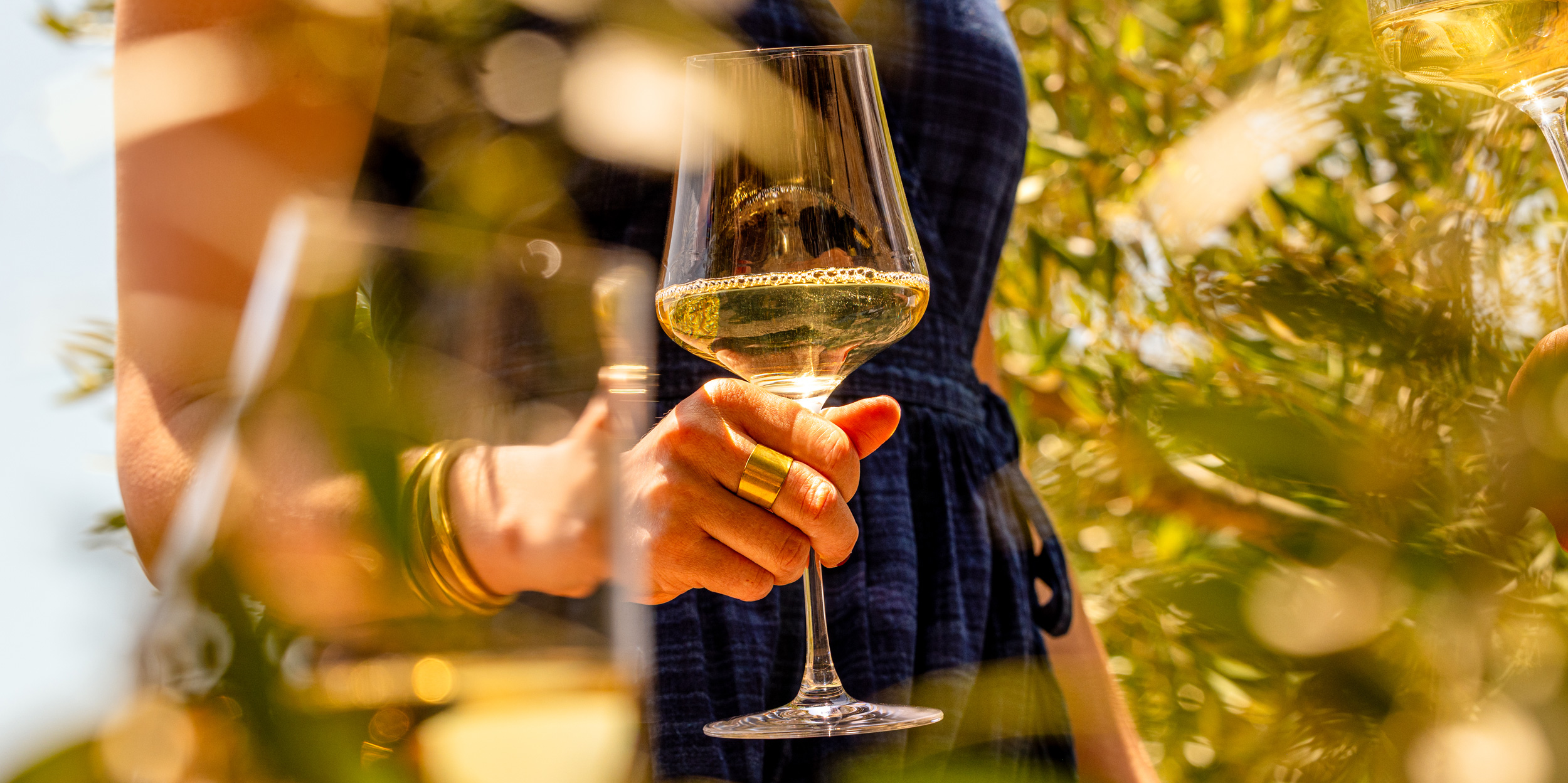 A woman in an olive grove, a glass of white wine in her hand. There is a second glass of white wine in the foreground.