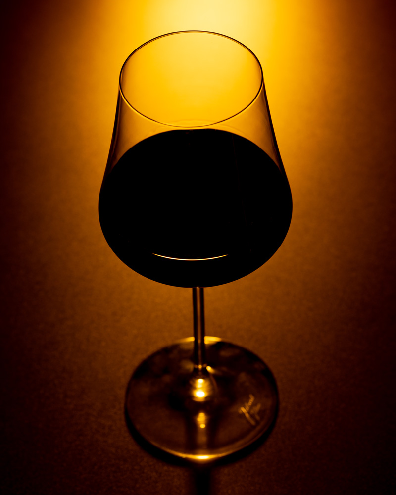 A silhouette of a wine glass filled with inky red wine, illuminated by a warm, yellow and orange light.